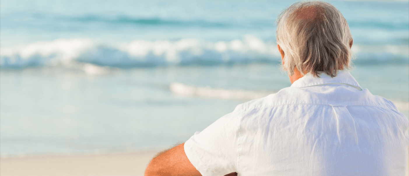 A silver-haired man in a white shirt seated on the sand, looking out over the surf
